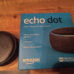 K&ouml;nnte beinhalten: Ein Amazon Echo Dot Smart Speaker und seine Box. Die Box ist t&uuml;rkis mit den Worten "echo dot" und "Add Alexa to any room". Die Box listet auch Sprachbefehle auf. Der Lautsprecher ist schwarz und rund.