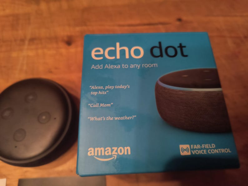 K&ouml;nnte beinhalten: Ein Amazon Echo Dot Smart Speaker und seine Box. Die Box ist t&uuml;rkis mit den Worten "echo dot" und "Add Alexa to any room". Die Box listet auch Sprachbefehle auf. Der Lautsprecher ist schwarz und rund.