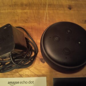 K&ouml;nnte beinhalten: Ein schwarzer Amazon Echo Dot Smart Speaker mit Netzteil und Kabel. Der Lautsprecher hat Lautst&auml;rke- und Stummschaltungstasten oben. Eine wei&szlig;e Karte mit "amazon echo dot" ist sichtbar.