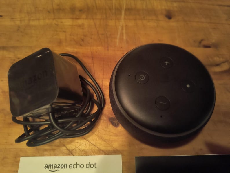 K&ouml;nnte beinhalten: Ein schwarzer Amazon Echo Dot Smart Speaker mit Netzteil und Kabel. Der Lautsprecher hat Lautst&auml;rke- und Stummschaltungstasten oben. Eine wei&szlig;e Karte mit "amazon echo dot" ist sichtbar.