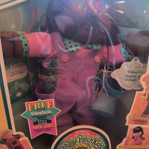 Könnte beinhalten: Cabbage Patch Kids Puppe in Originalverpackung. Die Puppe trägt eine rosa Latzhose, ein grün gepunktetes Hemd und hat braune Haare. Die Verpackung enthält Werbeaufkleber und das Cabbage Patch Kids Logo.
