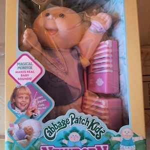 May include: A Cabbage Patch Kids Newborn doll in a box. The doll has a smiling face, wearing a purple and gray outfit. The box includes a pink monitor and text that reads "Magical Monitor Makes Real Baby Sounds!" and "Newborn with Magical Monitor."