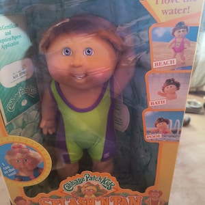 Könnte beinhalten: Cabbage Patch Kids Splash 'N Tan Puppe in Originalverpackung. Die Puppe hat braune Haare, blaue Augen und trägt einen grün-violetten Badeanzug. Auf der Box steht der Text "I love the water!" und Bilder der Puppe am Strand, im Bad und im Pool.
