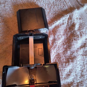 Könnte beinhalten: Geöffneter schwarzer Vintage-Kamera-Blitz mit leicht geöffnetem Deckel. Der Blitz hat eine kleine Glühbirne, zwei reflektierende Paneele und eine rote Basis. Die inneren Komponenten sind sichtbar, einschließlich eines weißen Papierstreifens und Metallteilen.