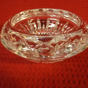 May include: A clear, round crystal bowl. It features a diamond-cut pattern around the rim and a starburst design in the centre. The bowl is displayed on a red surface, highlighting its intricate details and reflective qualities.