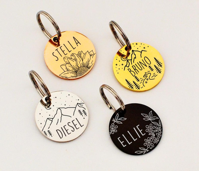 May include: Four round pet ID tags with silver key rings. The tags are gold, yellow, silver, and black. Each tag has a different design: a sunflower, mountains and trees, mountains and stars, and flowers. Each tag has a pet's name.