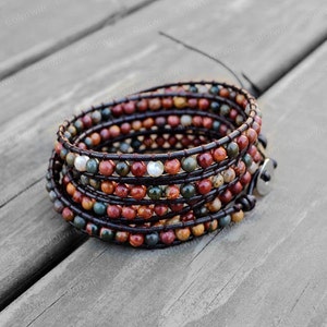 Puede incluir: Un brazalete de envoltura multi-cadena con cordón de cuero marrón y cuentas de piedras preciosas de colores. El brazalete tiene un cierre plateado y una perla blanca.