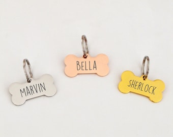 May include: Three dog bone-shaped name tags with rings. The tags are silver, rose gold, and gold. The names "Marvin", "Bella", and "Sherlock" are engraved on the tags.