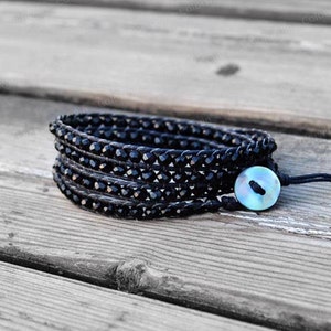 May include: A black beaded wrap bracelet with a blue and green button closure. The bracelet is made of multiple strands of beads and is wrapped around the wrist.