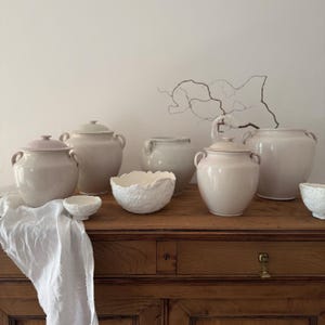 Op de afbeelding: Een verzameling keramische potten in verschillende vormen en maten, waaronder potten met deksel en kommen met textuur. De stukken zijn in crème- en lichtroze tinten en gerangschikt op een houten oppervlak. Afmetingen in centimeters.