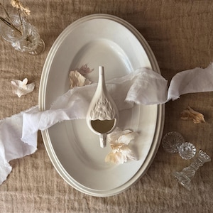 Peut inclure: Un plat de service ovale crème avec une pièce décorative en céramique en forme de goutte d'eau. La pièce en céramique présente un motif floral texturé. Du tissu blanc et des fleurs séchées sont disposés sur le plat.