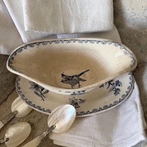 Könnte beinhalten: Eine cremefarbene Keramik-Sauciere mit blauem Blumenrand und einer Vogelabbildung in der Mitte, die auf einer passenden ovalen Untertasse steht. Mehrere Löffel mit schillernden Perlmutt-Laffen und verzierten silberfarbenen Metallgriffen liegen daneben.