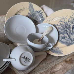 Peut inclure: Un ensemble de tasses et soucoupes en céramique blanche accompagné de deux assiettes crème ornées d'illustrations paysagères bleues. Une tasse est retournée, révélant la marque "Sarreguemines France" sur sa base. Une petite cuillère est posée dedans.