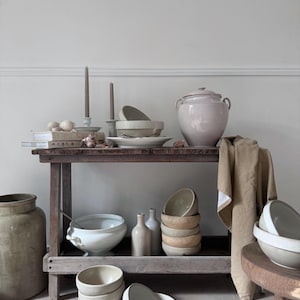 Op de afbeelding: Een rustieke houten tafel toont een verzameling keramische kommen, borden en potten in neutrale tinten. Twee lange kaarsen staan op de bovenste plank, naast een grote pot met deksel. Een linnen doek hangt aan de zijkant.