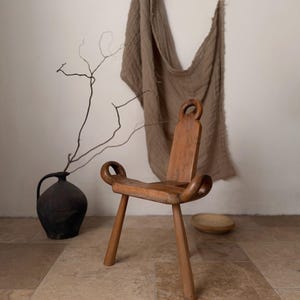 Può includere: Sedia in legno fatta a mano con un design unico. La sedia presenta una seduta curva, tre gambe e uno schienale con maniglia circolare. La venatura naturale del legno è visibile. Un elemento decorativo.