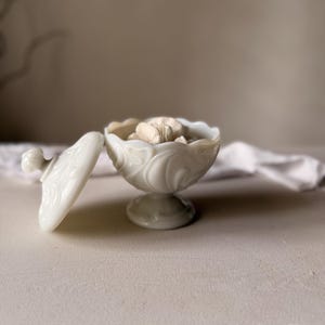 Może przedstawiać: Biała, nakrywana miseczka na nóżce z kwiatowym wzorem. Pokrywka jest zdjęta, odsłaniając beżowy przedmiot w środku. Miseczka ma falisty brzeg i stoi na okrągłej podstawie. Pokrywka ma mały, okrągły uchwyt. Miseczka jest prawdopodobnie wykonana z ceramiki lub szkła.