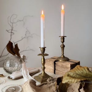 Può includere: Due candele bianche accese in candelabri antichi in ottone. I candelabri hanno basi e top dettagliati. Una pila di libri antichi ed elementi decorativi completano la natura morta.