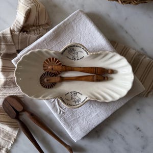 May include: An off-white, scalloped ceramic butter dish featuring a cow illustration and the text "BEURRE CHARENTES POITOU TRADITION DU GOUT." Two wooden pastry crimpers with fluted wheels rest inside the dish, and a pair of wooden tongs lies nearby.