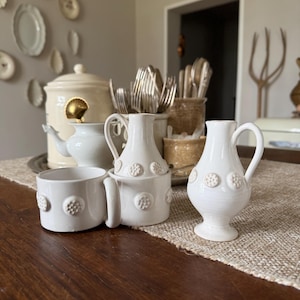 Puede incluir: Una colección de vajilla de cerámica blanca, que incluye una jarra pequeña, una taza y un jarrón decorativo, todos adornados con un patrón de puntos en relieve. Los artículos están dispuestos sobre una mesa de madera, lo que sugiere un entorno de cocina o comedor.