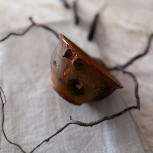 Può includere: Una piccola ciotola rotonda in terracotta marrone con interno smaltato e superficie esterna rustica e ruvida. La ciotola presenta due piccole maniglie sporgenti sui lati opposti e mostra variazioni di colore naturali nella finitura.