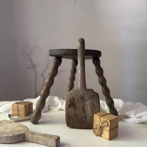 May include: A small, dark wooden stool with a round seat and three twisted legs. A large, weathered wooden scoop stands in front of the stool. Two small, wrapped packages and a wooden paddle are also present, suggesting a vintage or farmhouse theme.