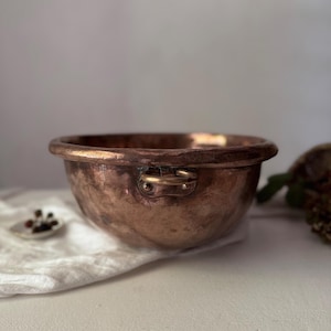 Peut inclure: Un bol en cuivre de forme arrondie avec deux petites poignées. Le bol présente une patine riche et vieillie, avec des variations visibles de couleur et de texture. Le bol est posé sur un tissu blanc.