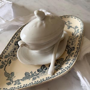 Könnte beinhalten: Eine weiße Keramik-Suppenterrine mit Deckel, Untertasse und Löffel, die auf einer cremefarbenen Servierplatte ruht. Die Platte hat ein blaues Blumenmuster an den Rändern.