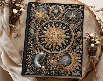 May include: Black journal with gold celestial designs, including a sun, moon, stars, and floral accents. The cover features intricate embossed details and colorful gemstones. The journal is placed on a wooden surface with dried flowers.
