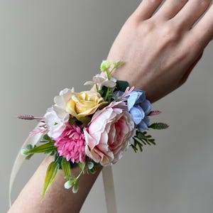 May include: A wrist corsage featuring a cluster of colorful artificial flowers. The arrangement includes pink, yellow, white, and blue blooms, accented with green foliage and cream-colored ribbons. A decorative floral accessory.