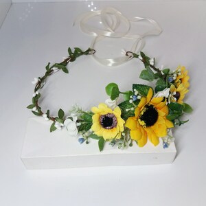May include: A sunflower and white flower crown with green leaves and a white ribbon.