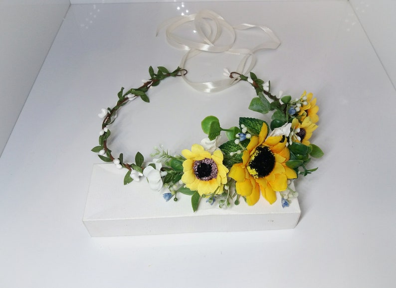 May include: A sunflower and white flower crown with green leaves and a white ribbon.