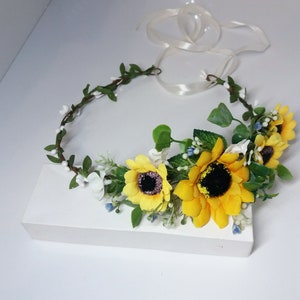 May include: A sunflower flower crown with yellow sunflowers, white flowers, and green leaves. The crown is tied with a white ribbon.
