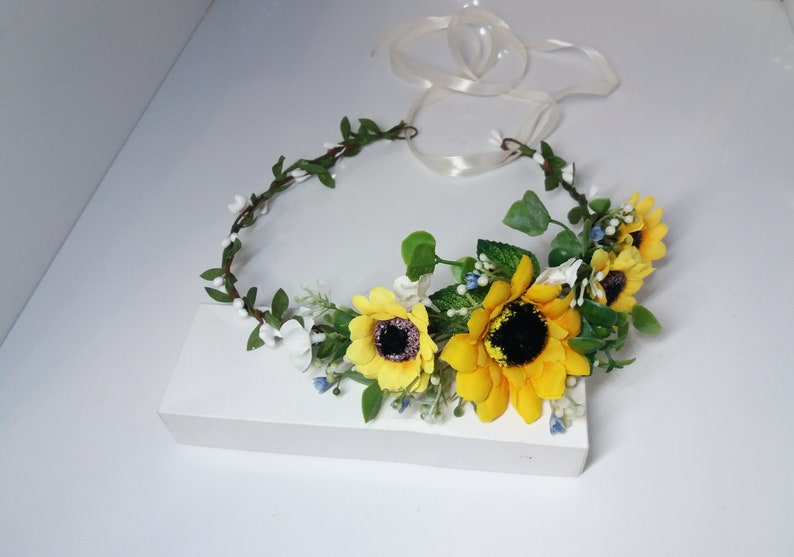 May include: A sunflower flower crown with yellow sunflowers, white flowers, and green leaves. The crown is tied with a white ribbon.
