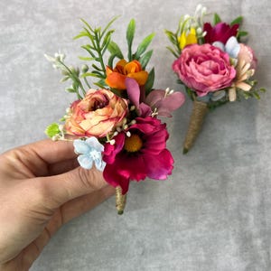 May include: Two floral boutonnieres with pink, orange, and yellow flowers. The boutonnieres are made with silk flowers and have a rustic burlap stem.
