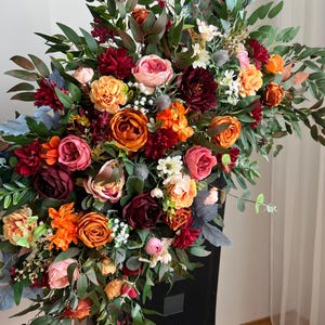 May include: A colourful floral display with a variety of flowers. The arrangement includes roses in shades of orange, pink, and burgundy, with green leaves. The flowers are arranged on a black chair, creating a striking contrast.