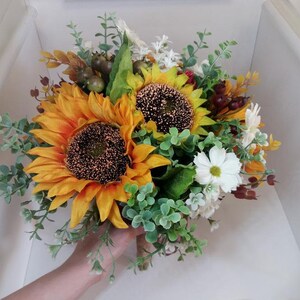 May include: A bridal bouquet featuring two large sunflowers with yellow petals and brown centers, surrounded by white daisies, green eucalyptus leaves, and small red berries.