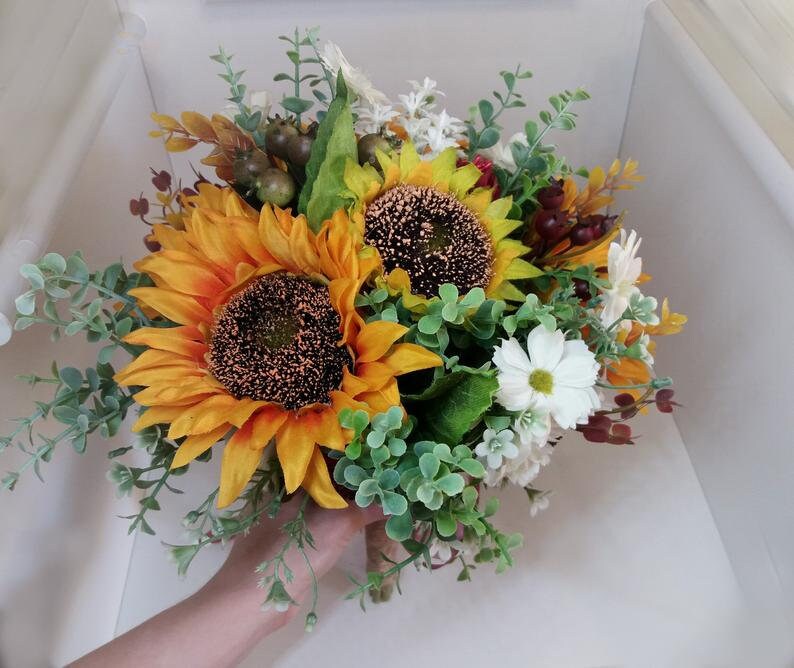 May include: A bridal bouquet featuring two large sunflowers with yellow petals and brown centers, surrounded by white daisies, green eucalyptus leaves, and small red berries.