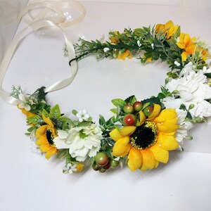 May include: A sunflower and white flower crown with green leaves and red berries. The crown is tied with a white satin ribbon.