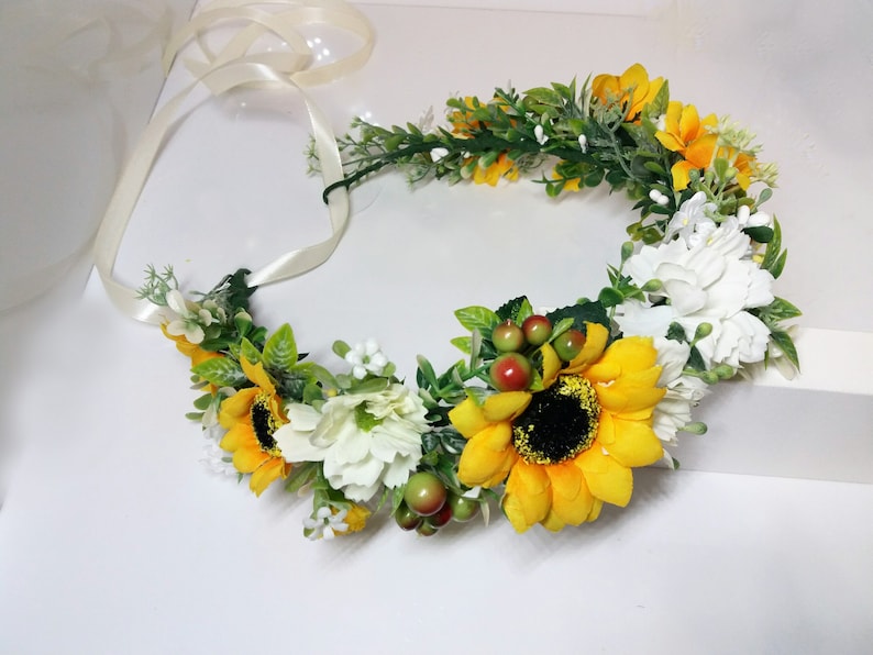 May include: A sunflower and white flower crown with green leaves and red berries. The crown is tied with a white satin ribbon.