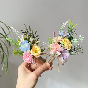 May include: Two floral boutonnieres with a variety of colorful flowers, including yellow roses, pink carnations, and blue blossoms. The stems are wrapped in twine. The arrangement is held in a hand.