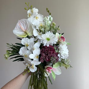Könnte beinhalten: Ein lebendiger Blumenstrauß mit weißen Orchideen, Gänseblümchen und Flamingoblumen, akzentuiert mit Grün und burgunderfarbenen Blüten. Das Arrangement wird in einer Hand gehalten und zeigt eine Vielzahl von Texturen und Farben, perfekt für ein Blumengeschenk oder die Inneneinrichtung.