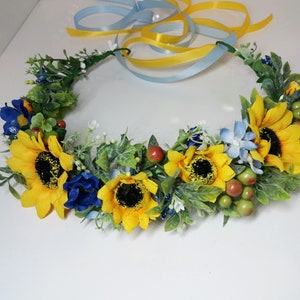 May include: A yellow sunflower and blue flower crown with green leaves and red berries.