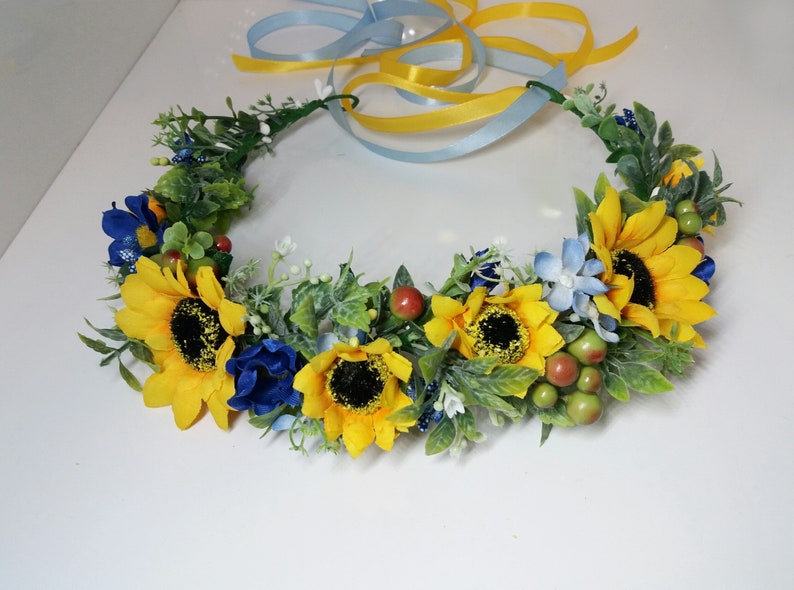 May include: A yellow sunflower and blue flower crown with green leaves and red berries.