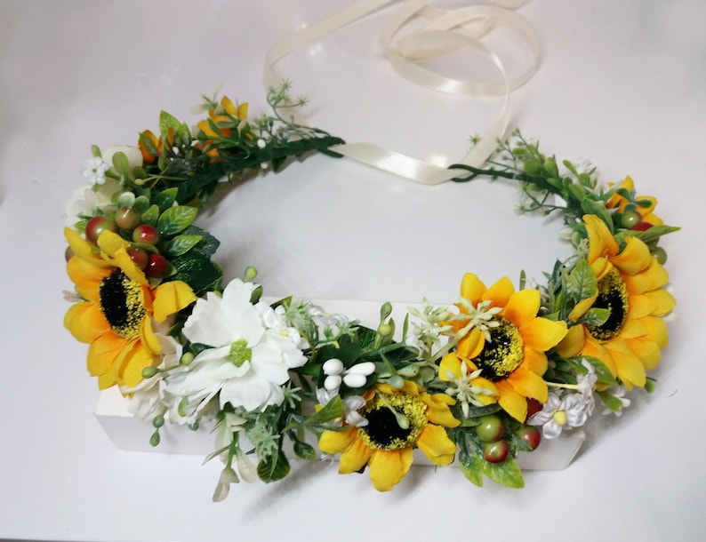 May include: A yellow sunflower flower crown with white flowers and green leaves. The crown is tied with a white ribbon.