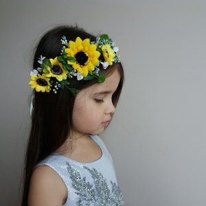 May include: A young girl wearing a headband with yellow sunflowers and blue and white flowers. The headband is made of green leaves and white flowers.