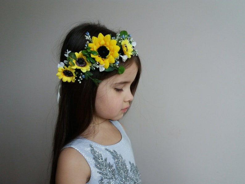 May include: A young girl wearing a headband with yellow sunflowers and blue and white flowers. The headband is made of green leaves and white flowers.