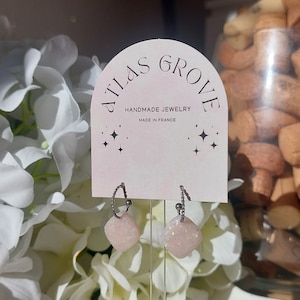以下が含まれることがあります： 銀製のフープピアスで、各フープに薄いピンク色のキラキラ光る石が取り付けられています。ピアスは、「Atlas Grove Handmade Jewelry Made in France」というテキストが書かれた白いカードに飾られています。