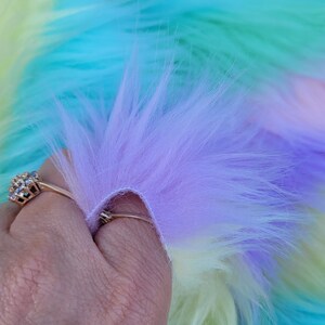 May include: A close-up of a multi-coloured faux fur fabric. The fabric is a gradient of light blue, yellow, and purple.