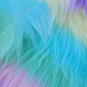 May include: A close-up of a rainbow-coloured faux fur fabric. The fabric is soft and fluffy, with a gradient of colours from purple to blue to green to yellow.