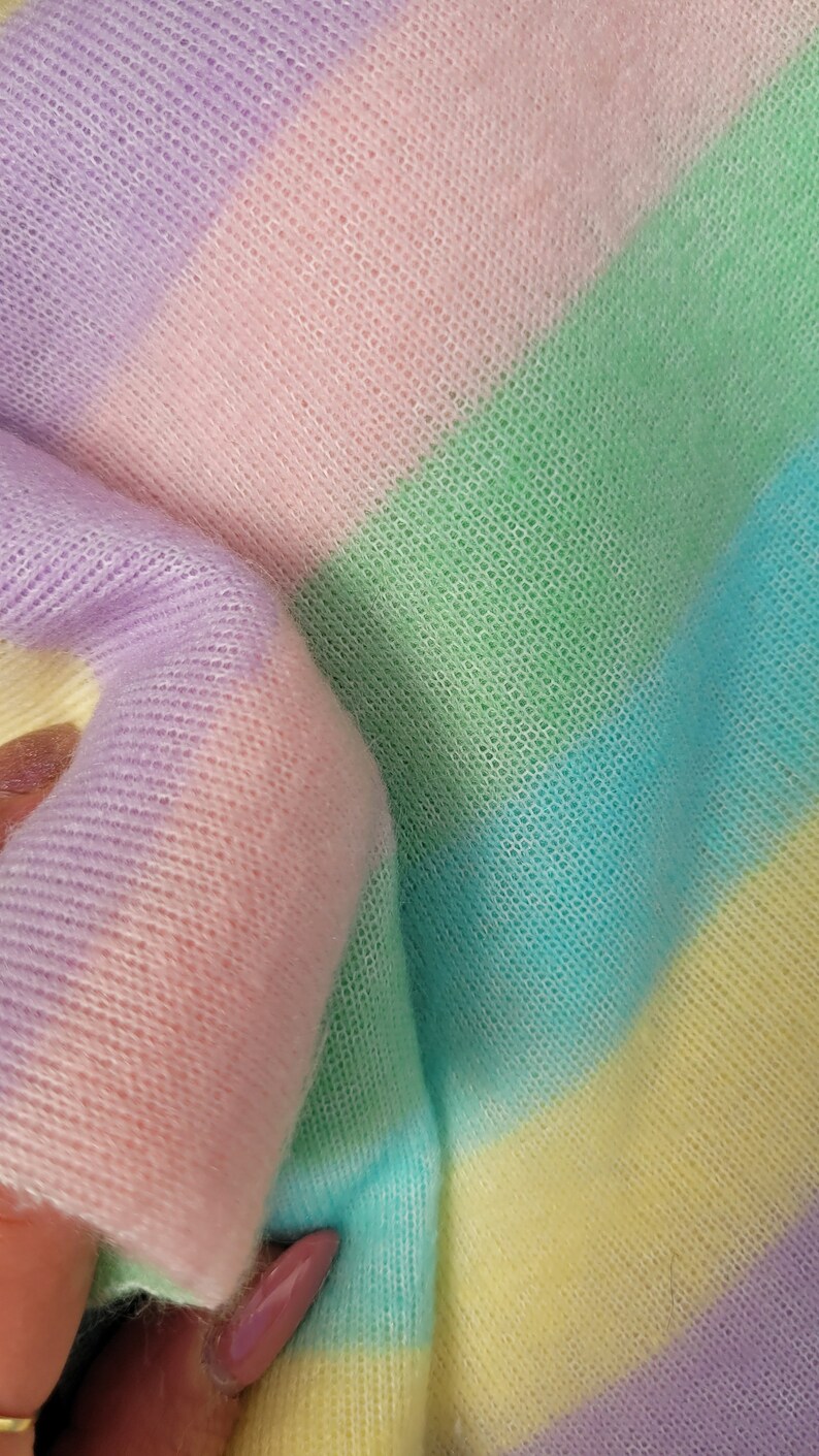 May include: A soft, striped knit fabric in pastel colours, including lavender, pink, green, blue, and yellow. The fabric is folded and shows the texture of the knit.